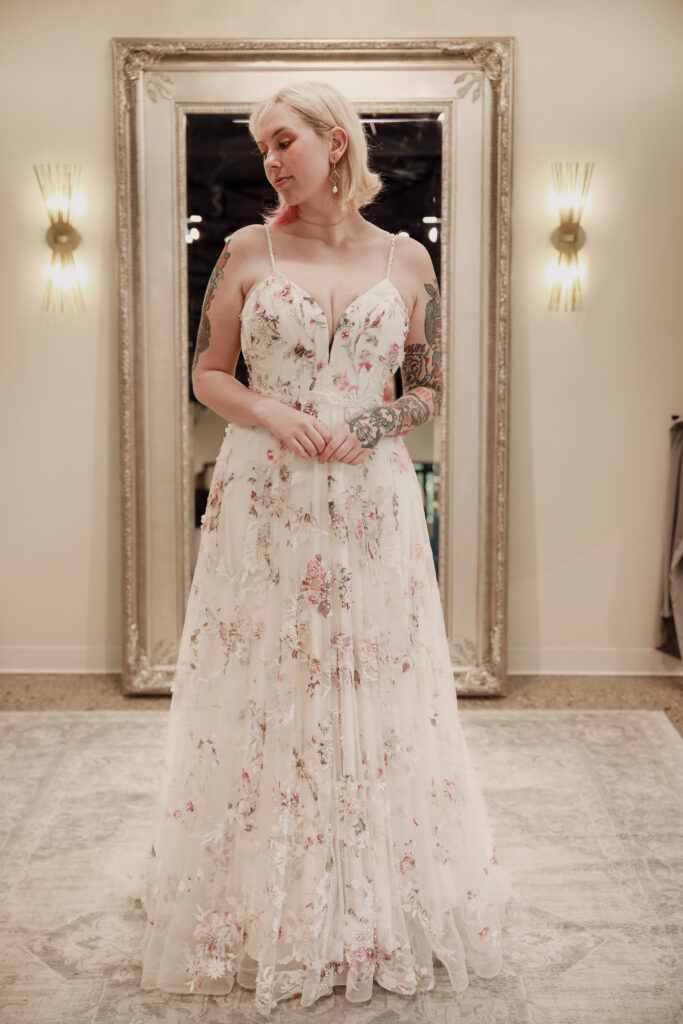 Bride in front of a mirror wearing a floral embroidered gown with a deep V-neckline in an elegant bridal suite.