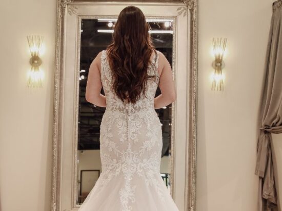 Rear view of a bride in a long white lace wedding dress, standing in front of a vintage mirror.