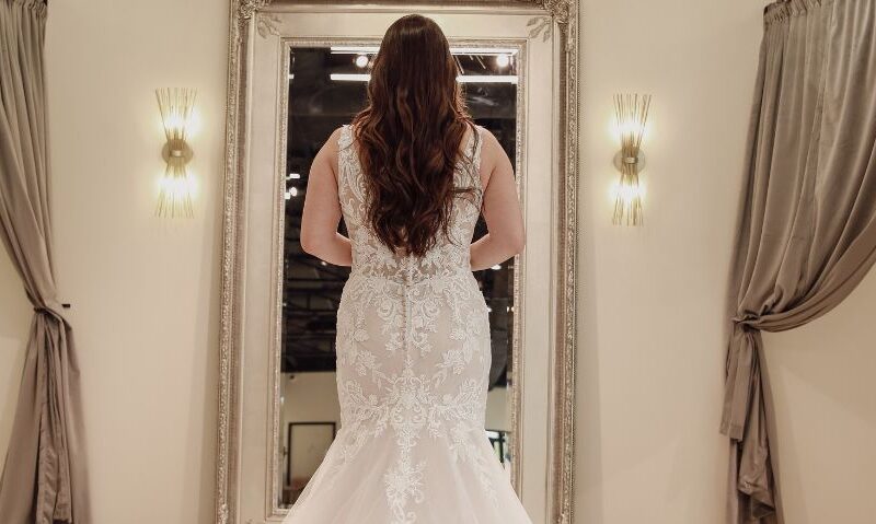 Rear view of a bride in a long white lace wedding dress, standing in front of a vintage mirror.