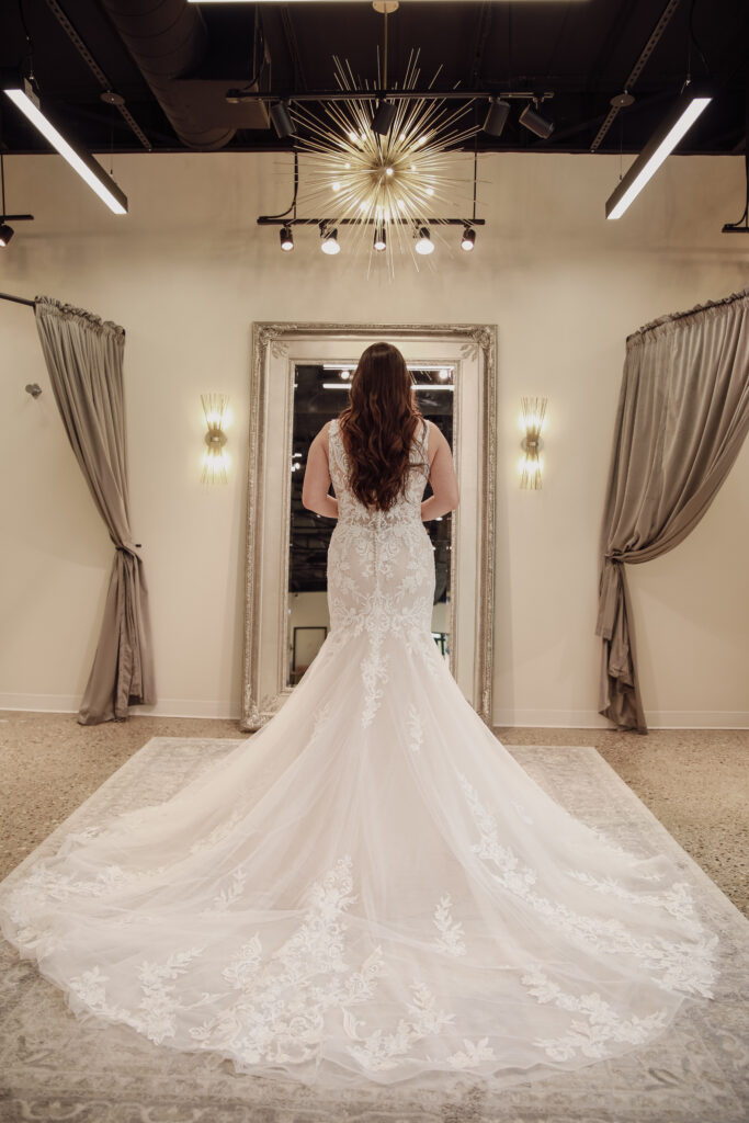 Bride in a white lace gown with a detailed train, facing a mirror in a well-lit bridal shop.