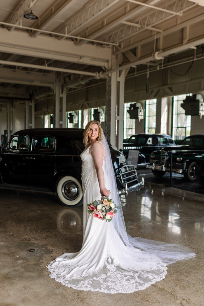 A bride posing in front of a classic automobile holding a pink and white flower bouquet by her side.