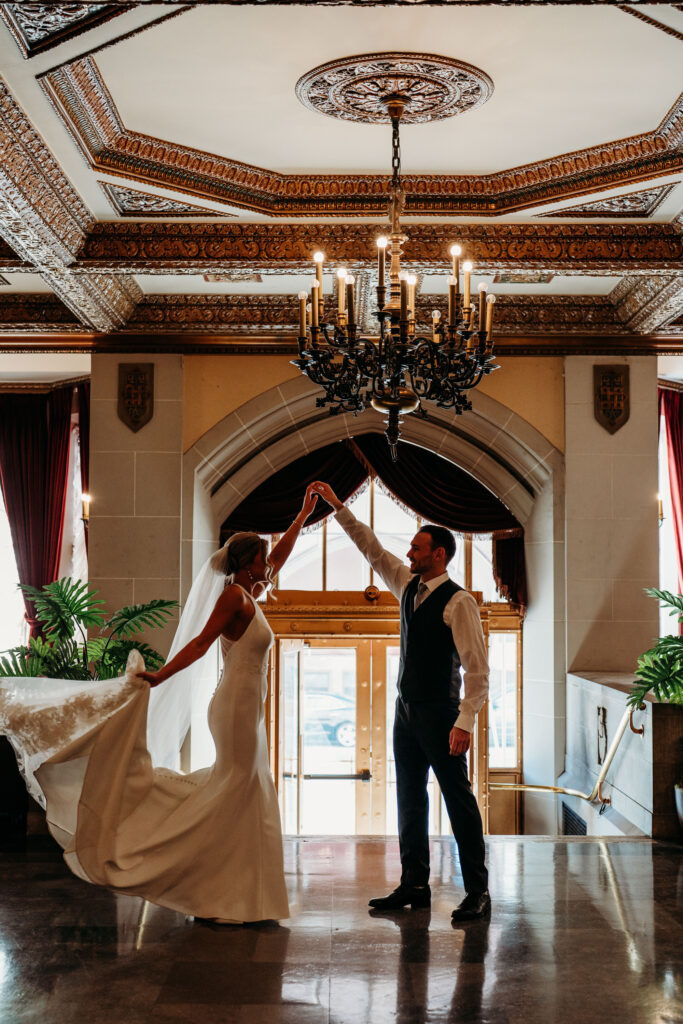 A bride and groom share a dance in an opulent venue with a grand chandelier and ornate ceiling details.