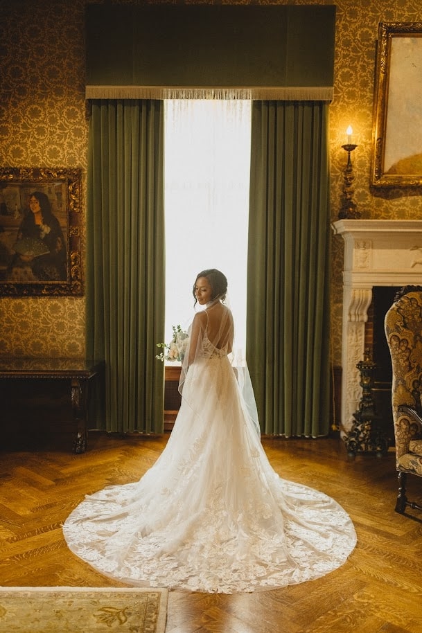 A bride stands gracefully in front of a large window with green curtains in a beautifully decorated room, showcasing the intricate lace details and long train of her wedding dress.