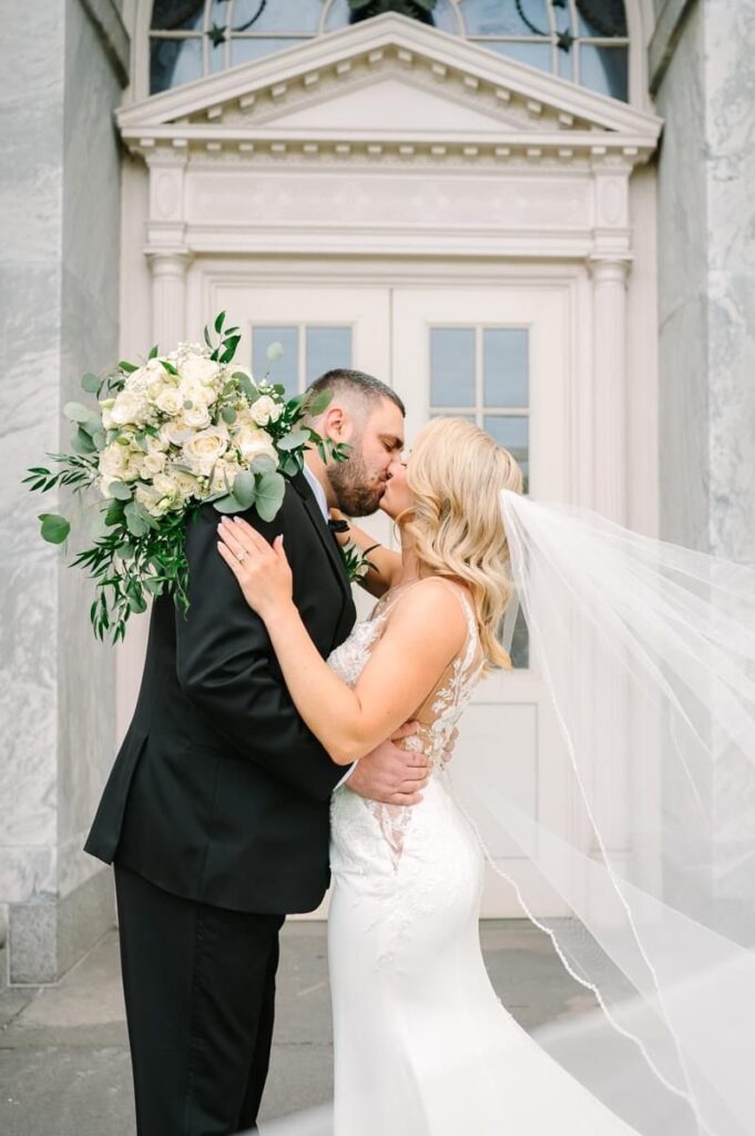 A couple kissing each other at their wedding venue as the bride holds a large bouquet of flowers.