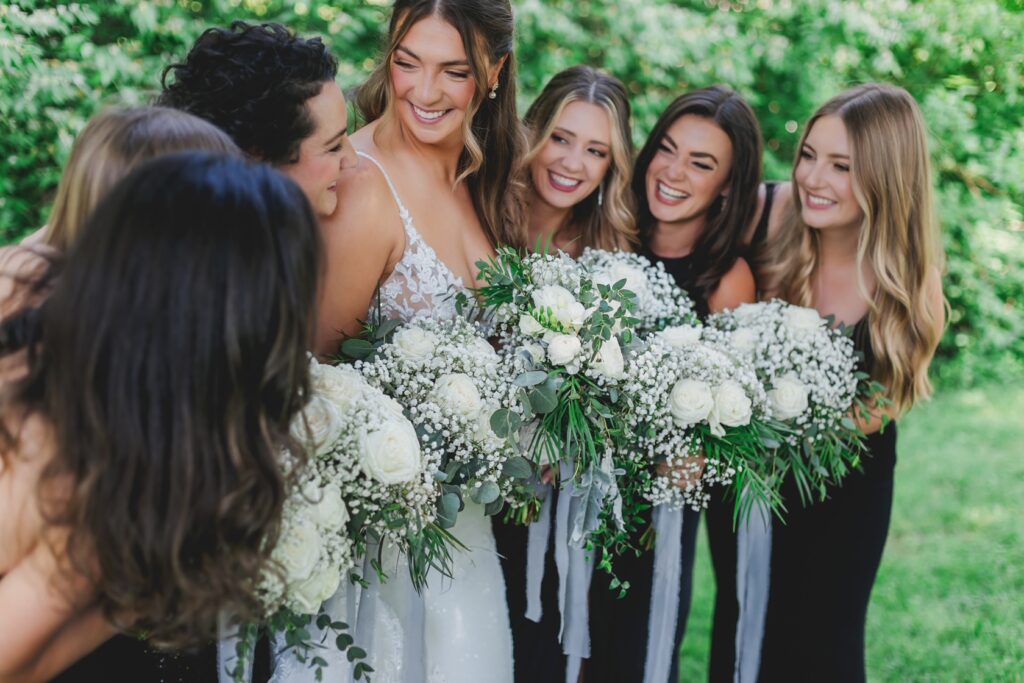 A group of smiling bridesmaids around the bride, each holding a bouquet of white flowers with green details.