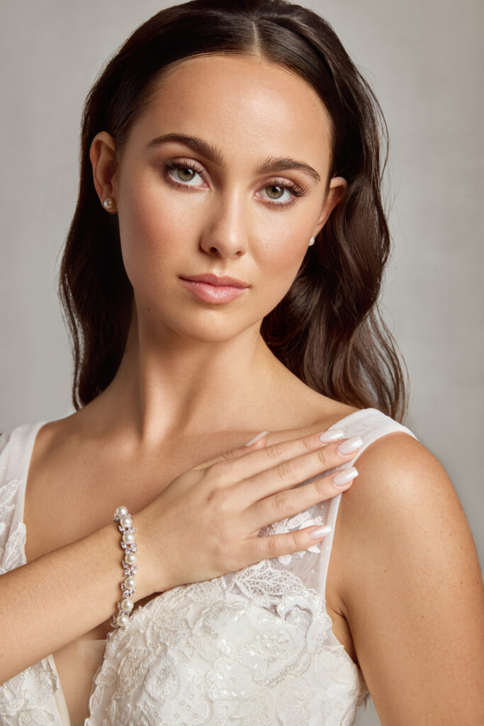 A close-up of a bride wearing a delicate pearl bracelet and a lace wedding dress with intricate floral details.