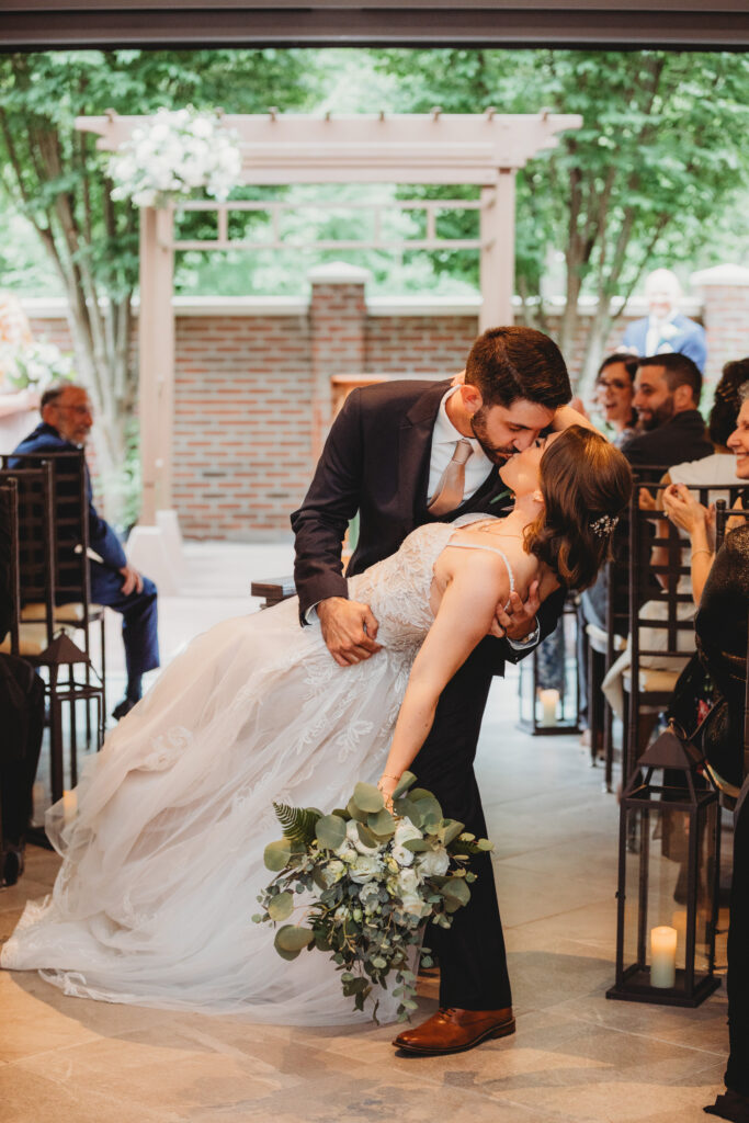 Bride dramatically dipped by groom during a kiss, surrounded by greenery.