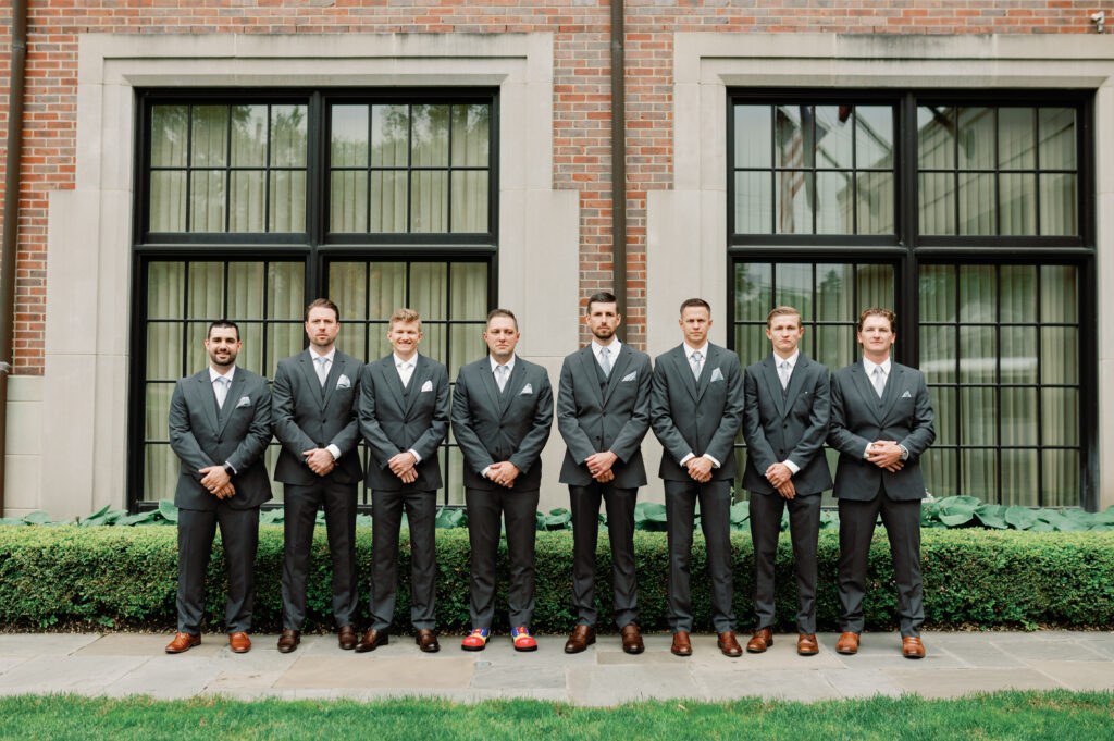 Groomsmen lined up in front of a building, all in grey suits with a garden backdrop.