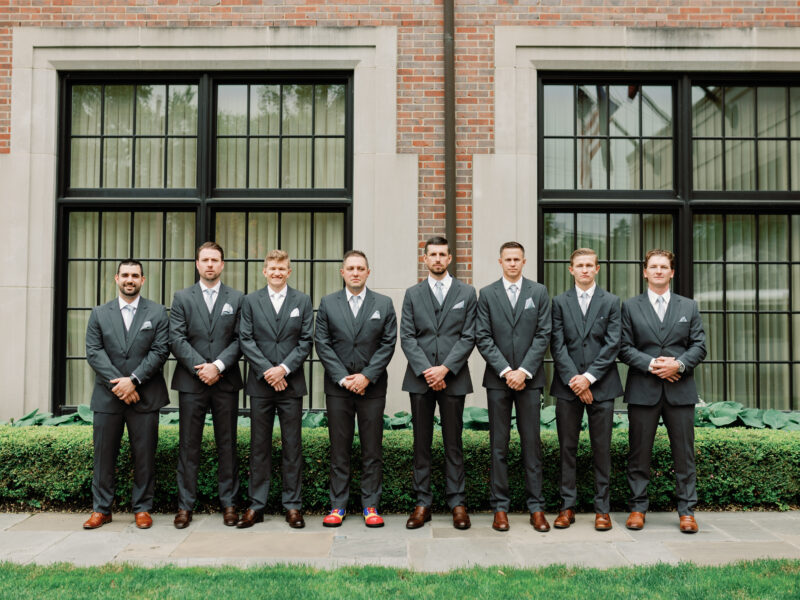 Groomsmen lined up in front of a building, all in grey suits with a garden backdrop.