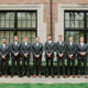 Groomsmen lined up in front of a building, all in grey suits with a garden backdrop.