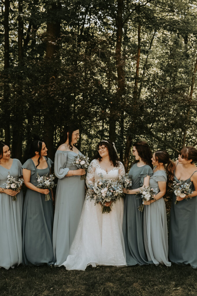 Bride with bridesmaids in blue dresses, all holding bouquets in a forest setting.