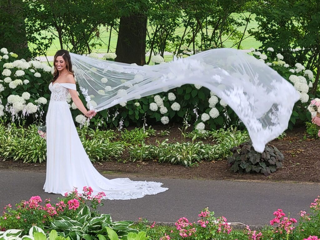 Bride playfully twirling her long, lace-trimmed veil among vibrant garden blooms.