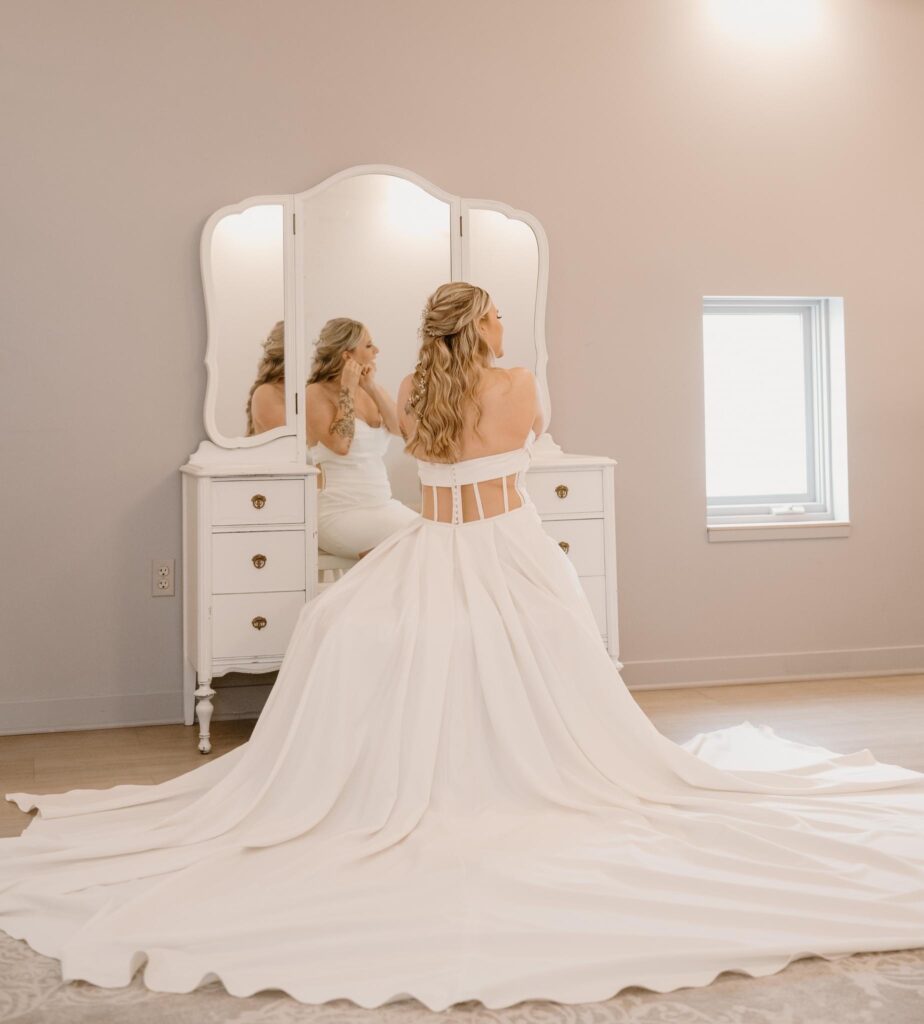 A bride putting on a pair of earrings while wearing a beautiful wedding dress in front of a mirror showing her reflection.