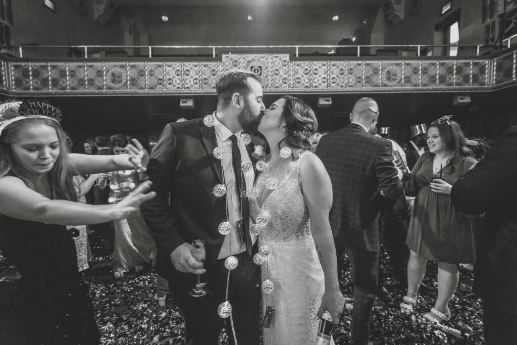 A bride and groom share a kiss while surrounded by guests at their wedding reception.