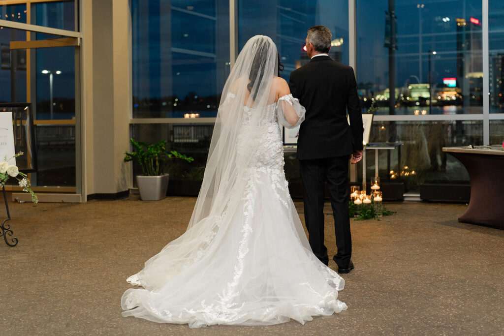 A bride and groom stand together, facing away from the camera, with the bride wearing an elegant lace wedding dress and a long veil.