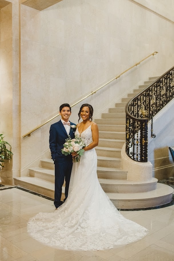 A bride and groom pose on a grand staircase in a luxurious venue. The bride wears a lace wedding dress with a long train, and the groom is dressed in a dark suit.