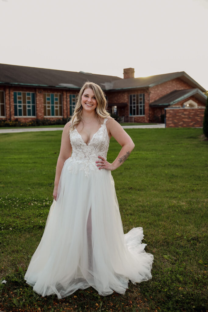 Smiling bride outdoors in a lace gown with a front slit, greenery in the background.