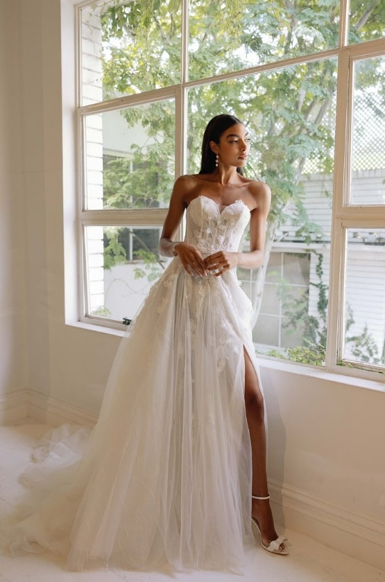 Stylish bride in a detailed lace gown with a long slit, standing in a bright room.