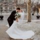 Bride and groom kissing under snowfall, surrounded by illuminated trees.