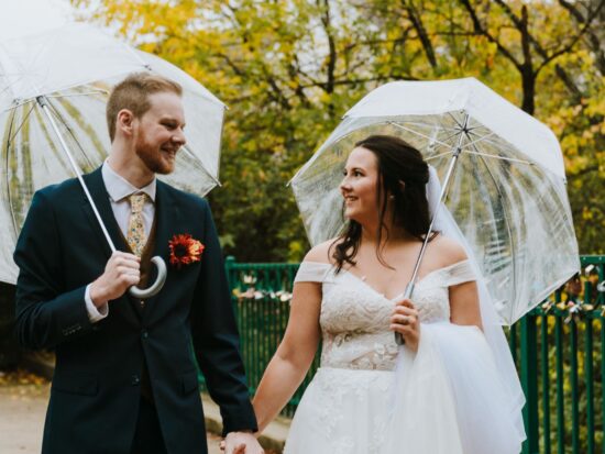 Joyful bride and groom under clear umbrellas on a leaf-strewn park bridge.