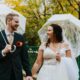 Joyful bride and groom under clear umbrellas on a leaf-strewn park bridge.