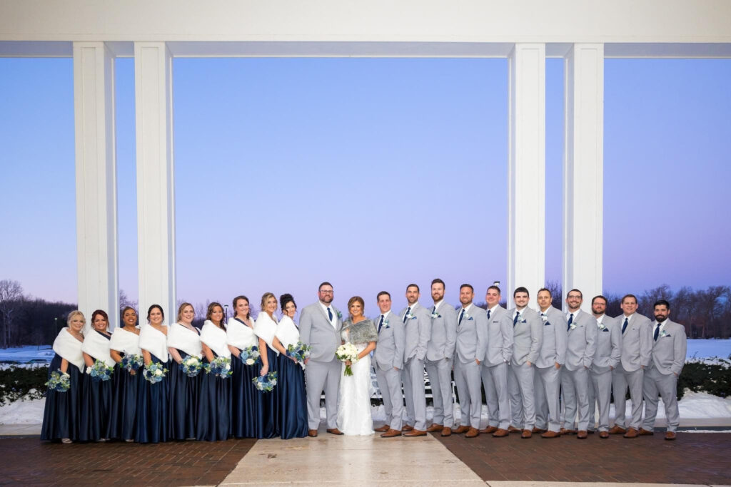 Large wedding party in blue and grey, posing under a large white pergola.