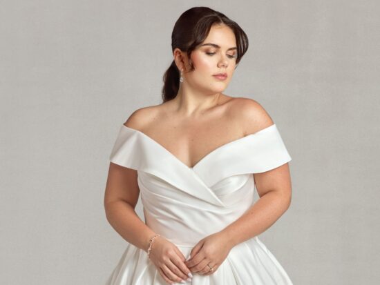 Bride introspectively looking down while standing in a sleek, off-shoulder white gown.