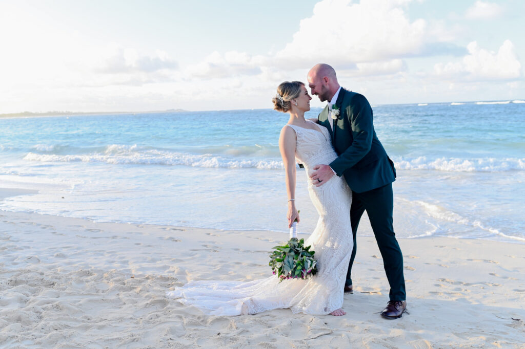 Bride and groom holding hands on a sandy beach, ocean waves lapping at the shore.