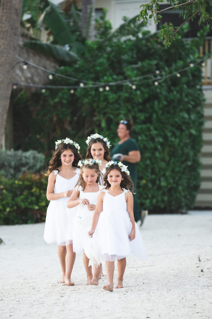 Four young flower girls walking on a sandy beach, wearing white dresses and floral crowns.