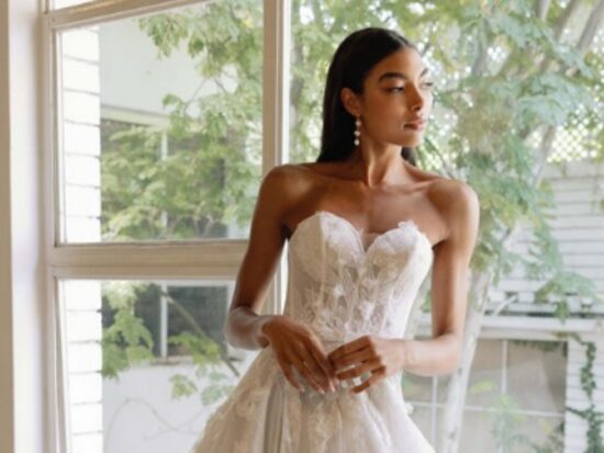 Elegant bride in a strapless lace gown with a high slit standing by a window.