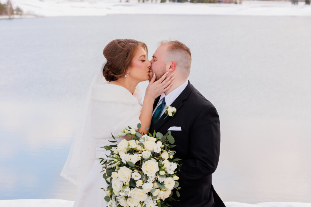 Bride and groom kissing by a frozen lake, bride in a white shawl.