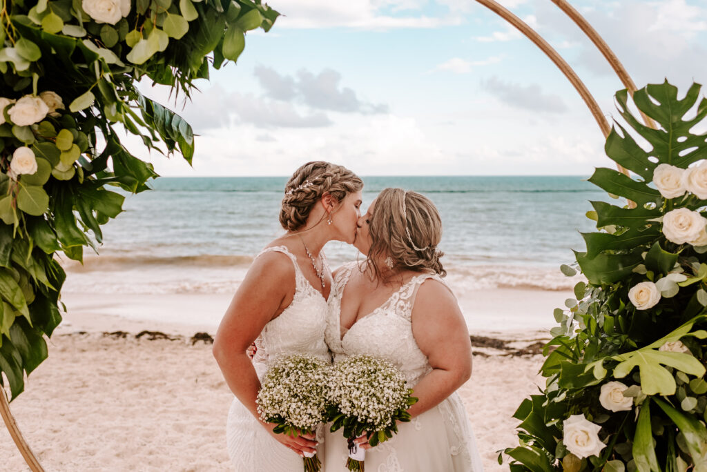 Two brides kissing on a tropical beach, framed by floral arches and ocean backdrop.