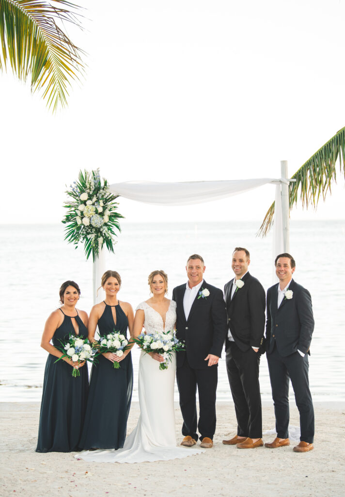 Wedding party by the sea, with bridesmaids in navy dresses and groomsmen in black suits.