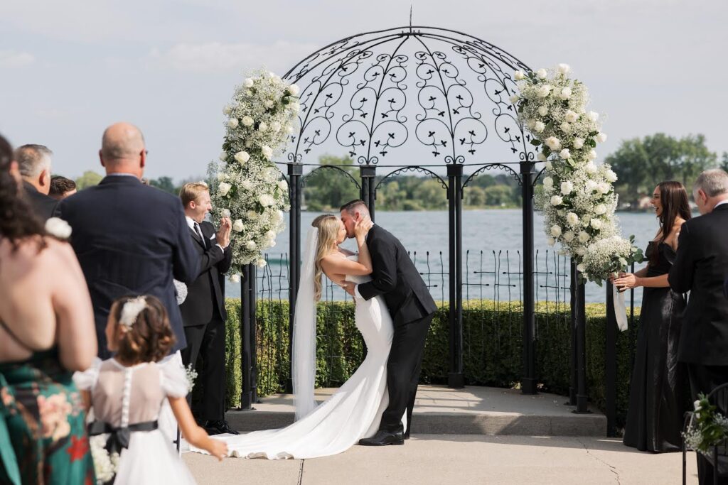 Newlyweds share a kiss under an intricate black metal arch surrounded by white flowers and a lake.