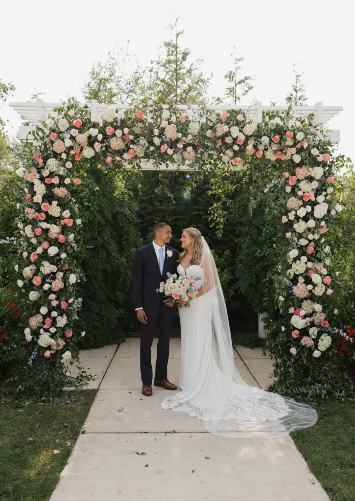 Bride and groom share a moment under a lavishly decorated floral arch in a lush garden setting.