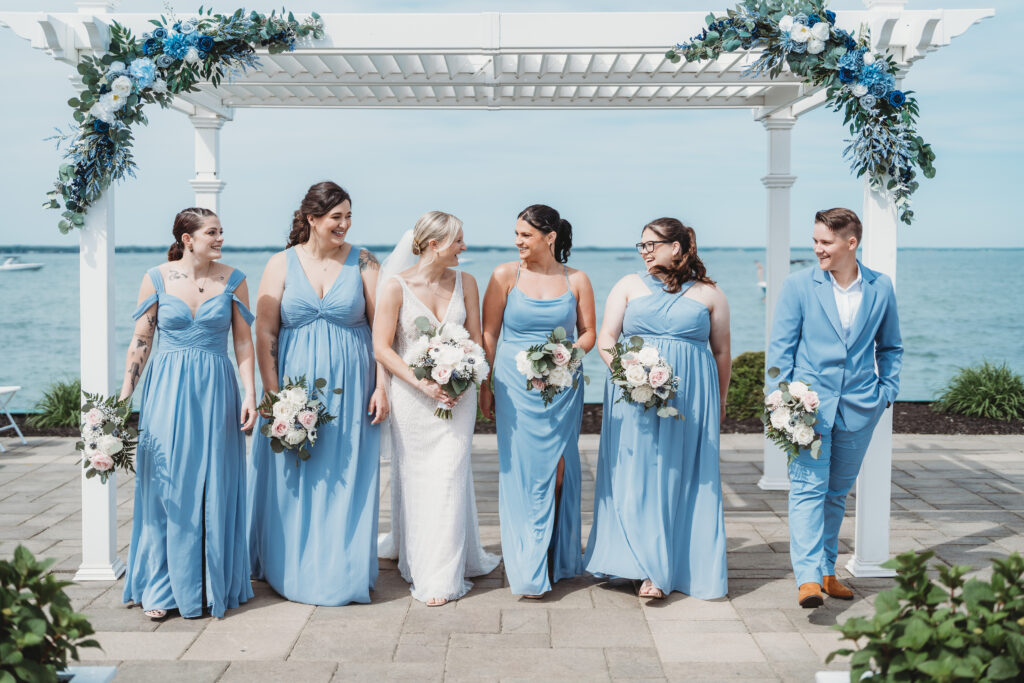 Wedding party in sky blue dresses smiles together under a floral adorned pergola by the lake.