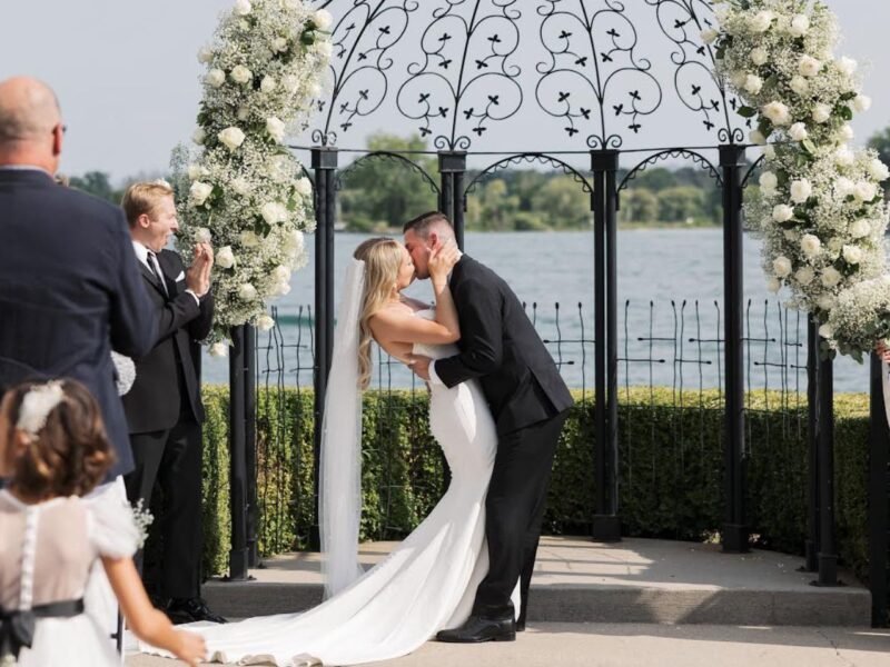 Bride and groom kiss passionately at a lakeside ceremony with an ornate floral arch backdrop.