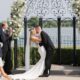 Bride and groom kiss passionately at a lakeside ceremony with an ornate floral arch backdrop.