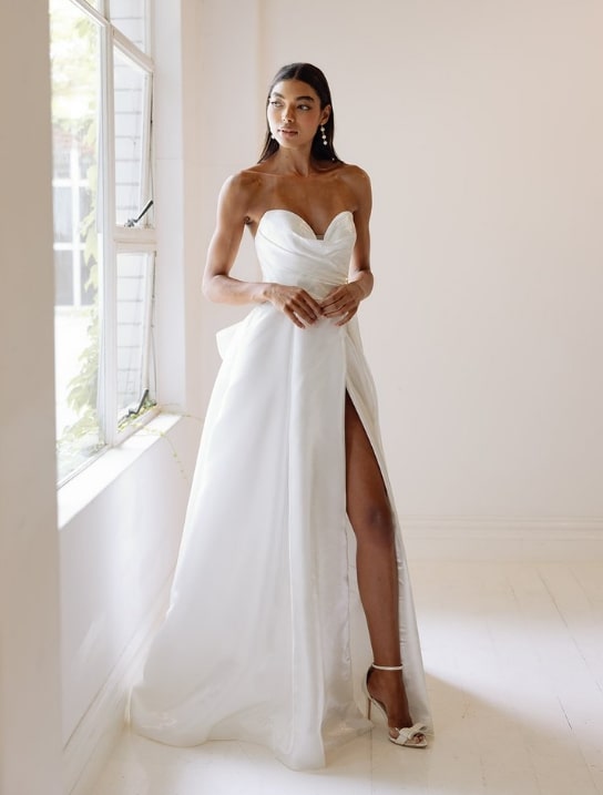 Sophisticated bride in a strapless gown with a high slit, posing beside a window.