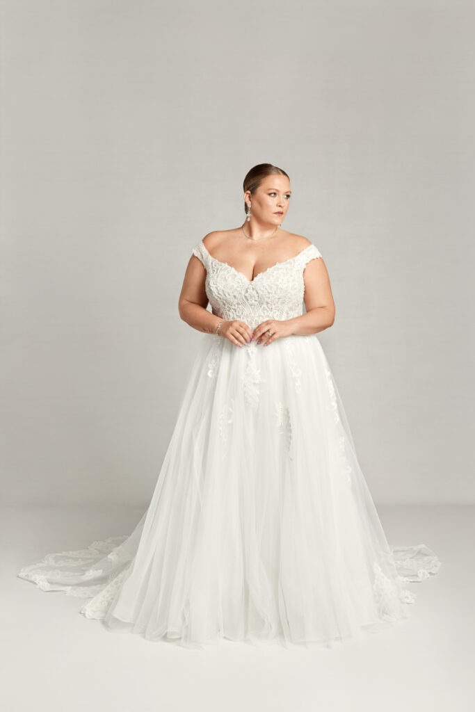 Bride in a white off-shoulder gown with delicate floral lace, posing regally.