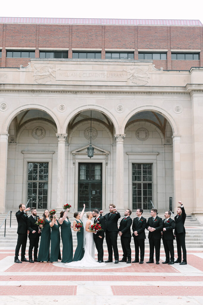 Bridal party celebrating with joy on the steps of William L. Clements Library, under clear skies.