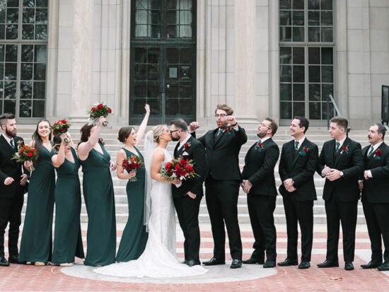 Bridal party celebrating with joy on the steps of William L. Clements Library, under clear skies.