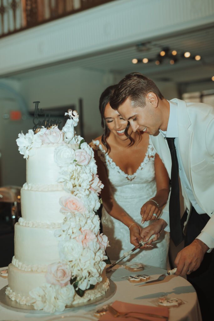 Bride and groom cutting into a tall white wedding cake adorned with white and blush floral accents and a custom topper.