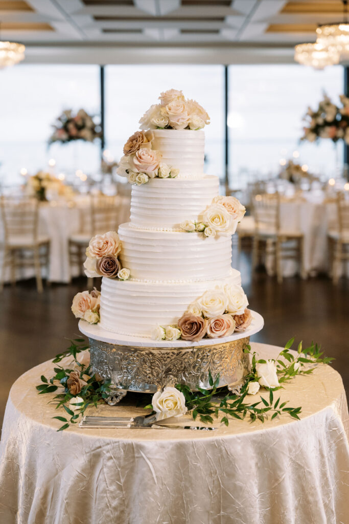 Elegant four-tier white wedding cake decorated with soft blush and ivory roses, displayed on a silver cake stand.