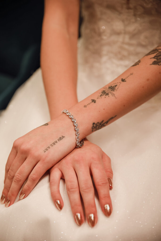 Hands with tattoos wearing a glistening diamond bracelet, resting on a wedding gown, with metallic nail polish adding contrast.