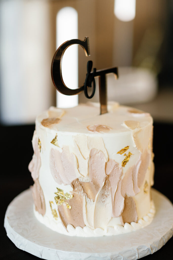 Small single-tier wedding cake with textured buttercream strokes in blush, ivory, and gold, featuring a metallic "C & T" cake topper.