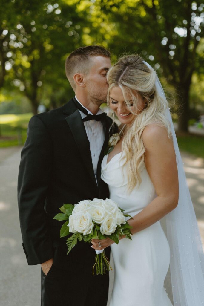 Bride and groom embrace outdoors, with the groom gently kissing the bride's forehead. Bride holds a bouquet of white roses.