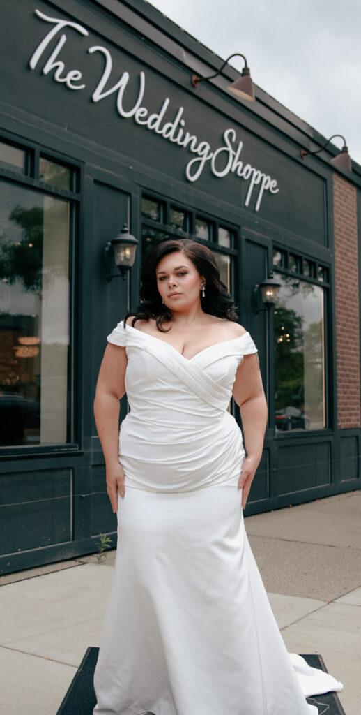 Plus-size bride in a classic white gown, posing outside The Wedding Shoppe, showcasing elegance and beauty.