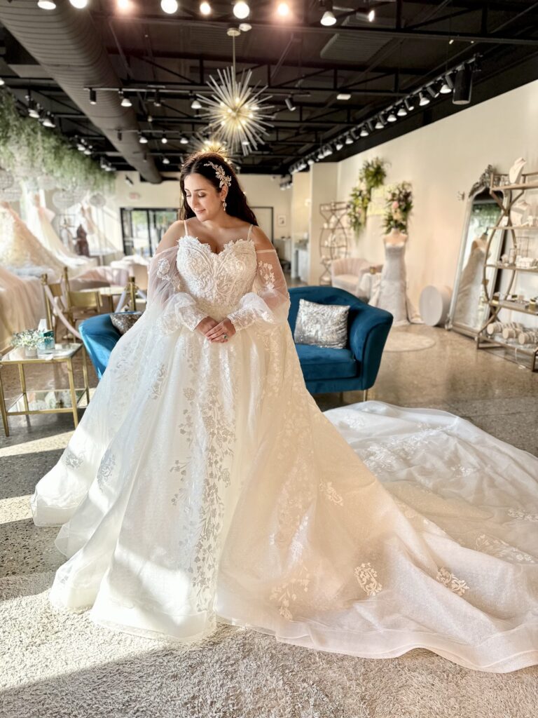 Plus-size bride in a flowing lace gown with a dramatic train, standing in a beautifully decorated bridal shop.