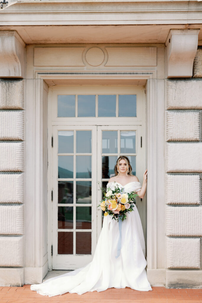 Bride in off-shoulder wedding dress stands framed by tall French doors, holding a lush bouquet of mixed flowers.